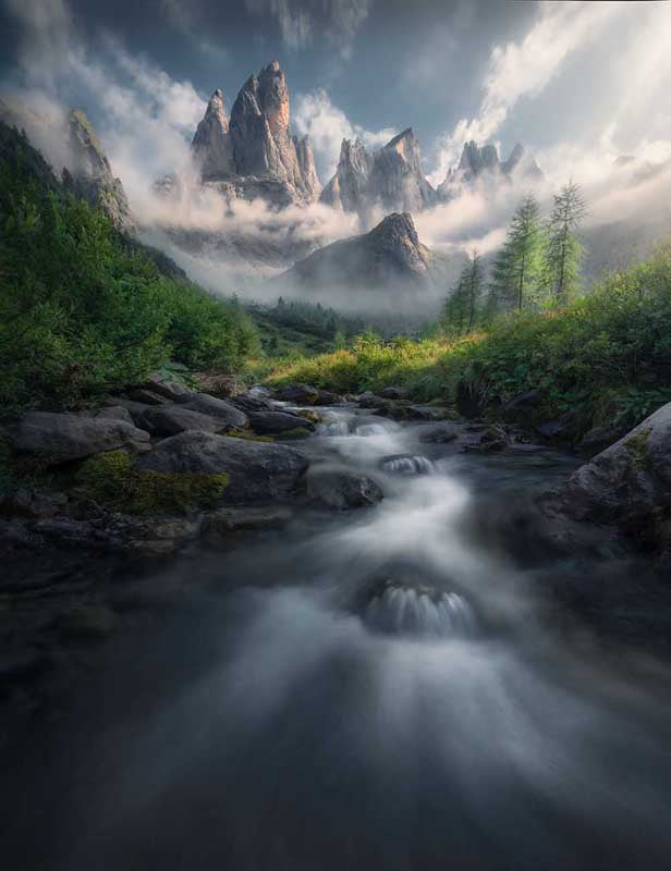 The mountains of the Dolomites in summer as seen on a misty morning with a stream flowing through a green valley of lush trees.