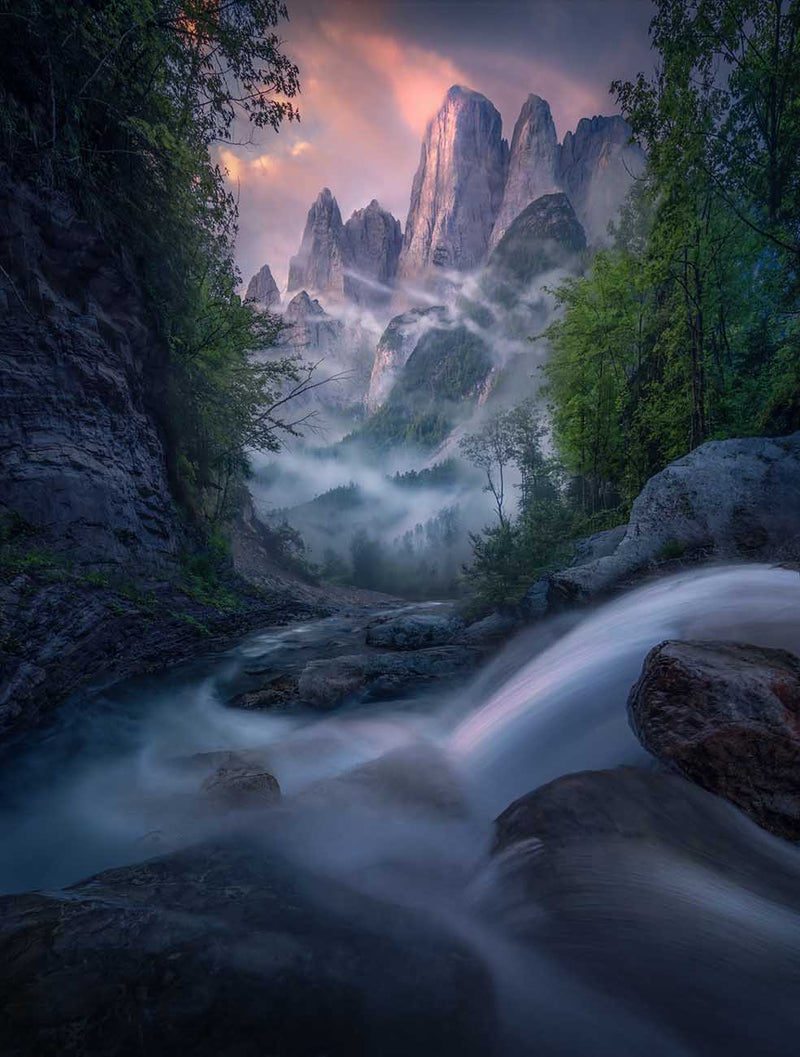 A majestic mountain landscape in the Dolomites in summer with a waterfall cascading towards the mountain peaks framed by tall green trees on each side. The clouds glows pink and mist streams through the valley and around the peaks.