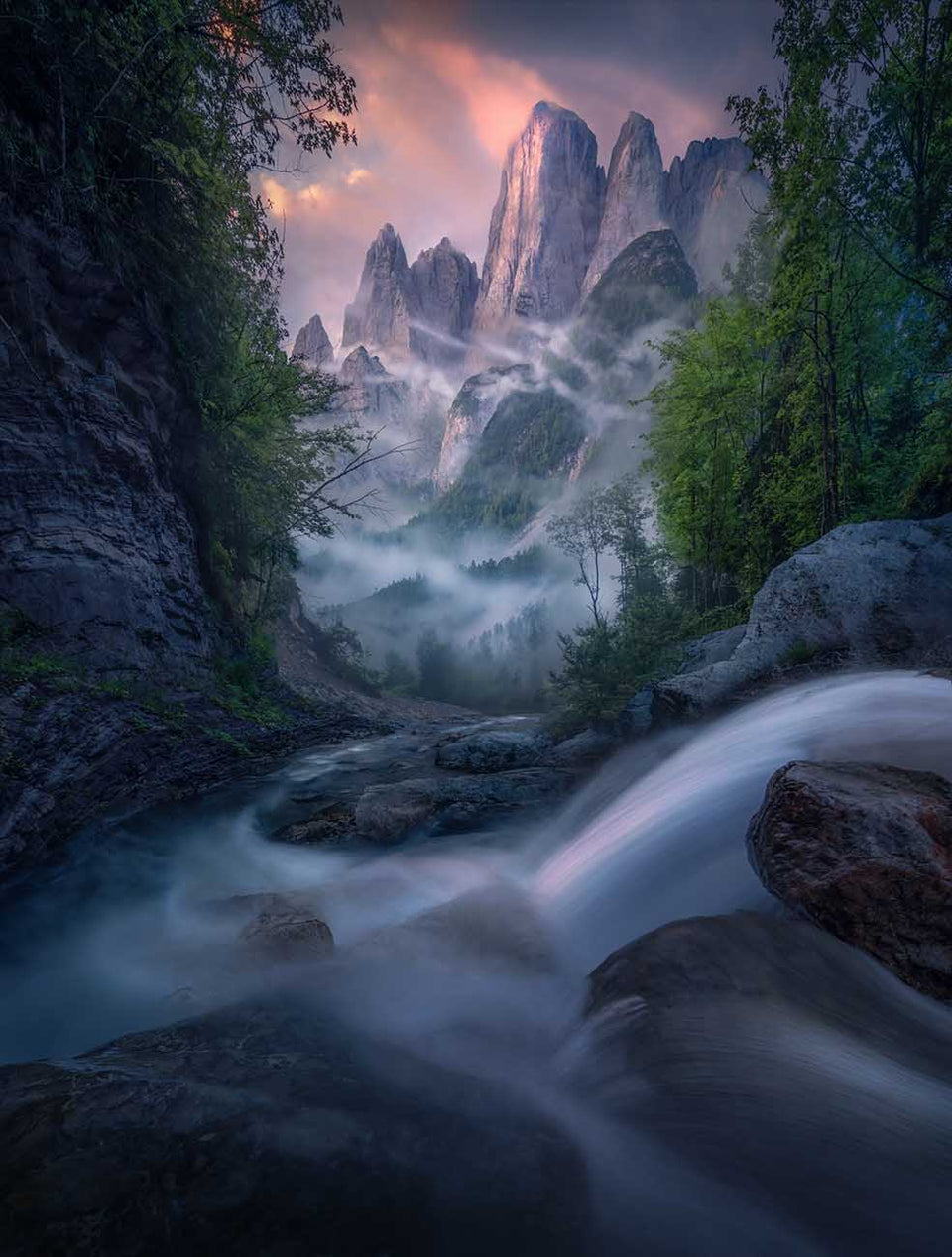 A majestic mountain landscape in the Dolomites in summer with a waterfall cascading towards the mountain peaks framed by tall green trees on each side. The clouds glows pink and mist streams through the valley and around the peaks.
