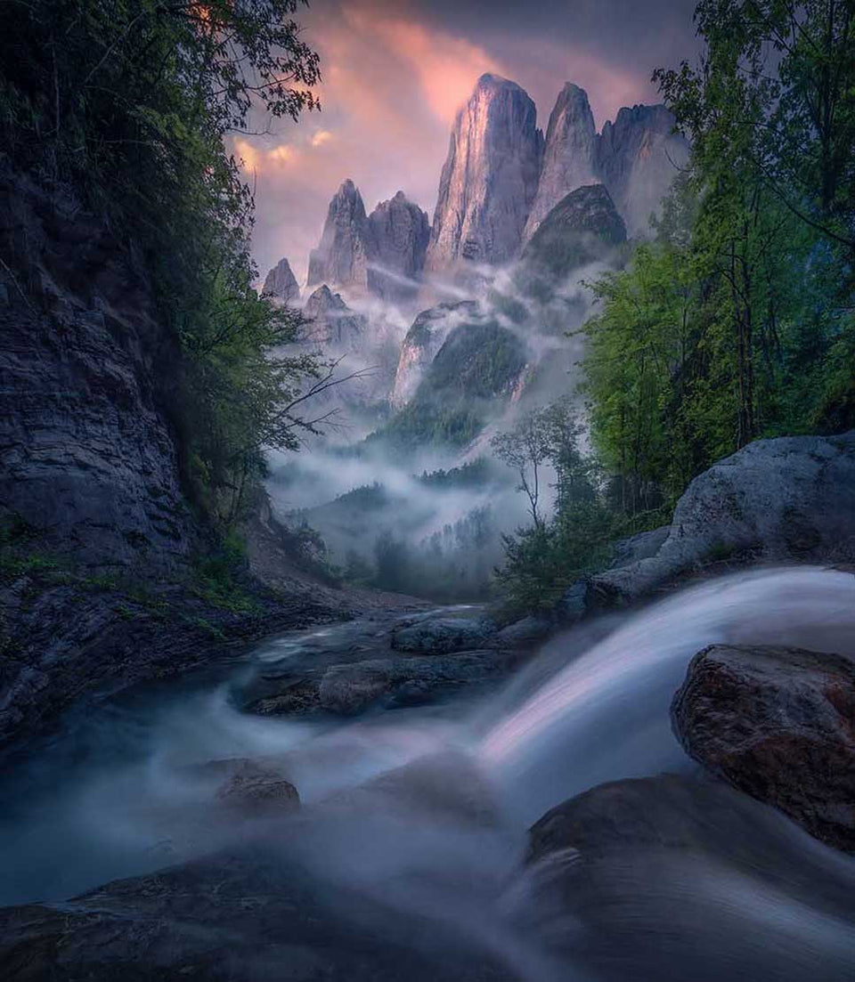 Impressive mountain faces shrouded with mist stand tall with a pink sunrise catching the sides of the mountains and the clouds. The peaks are framed by forest trees and in the foreground a waterfall winds through the forest to the peaks.