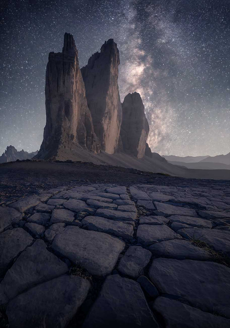 A dramatic night landscape photograph of Tre Cime, the three tall rock spires under a star-filled sky with the Milky Way, with cracked dark ground in the foreground.