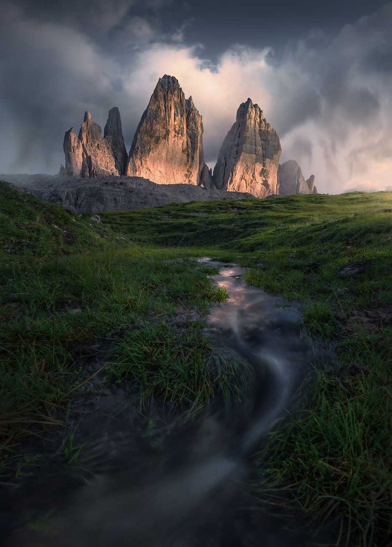 Drei Zinnen in the Dolomites, the three jagged rock spires rising above a grassy field, with a small stream flowing through the foreground under a dark, moody sky with light breaking through the clouds.
