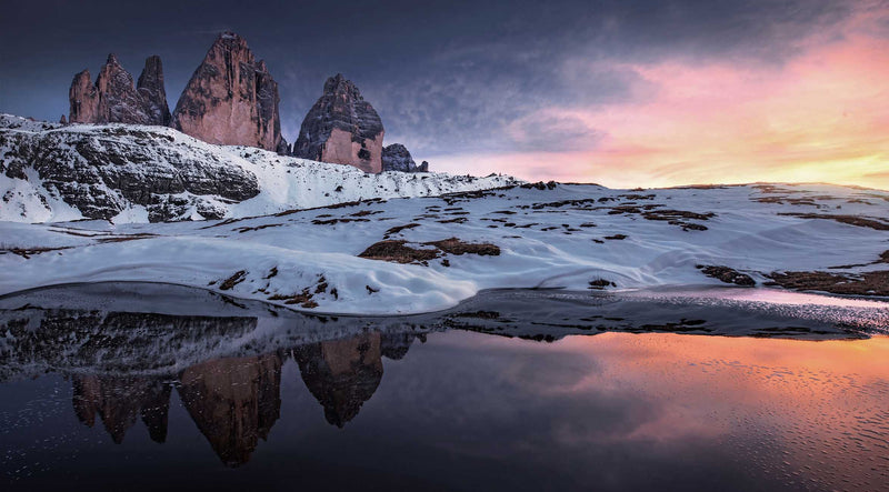 The Drei Zinnen three tall rock spires in winter snow-covered mountains reflected in a calm lake under a pink, orange, and blue sunset sky.