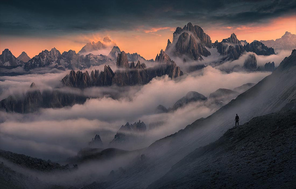 Taken from Drei Zinnen in the dolomites. Layers of mountains can be seen through the misty clouds and pink and orange sunset colours light the sky. A silhouette is seen viewing the mountains from the right.
