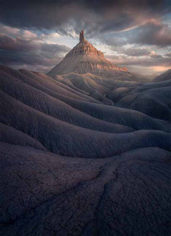 Factory Butte at sunset glows a pink orange, the sky is tinted the same colours. in the foreground deep channels of mud hardened by rain and layers of sediment form layers of small canyons.