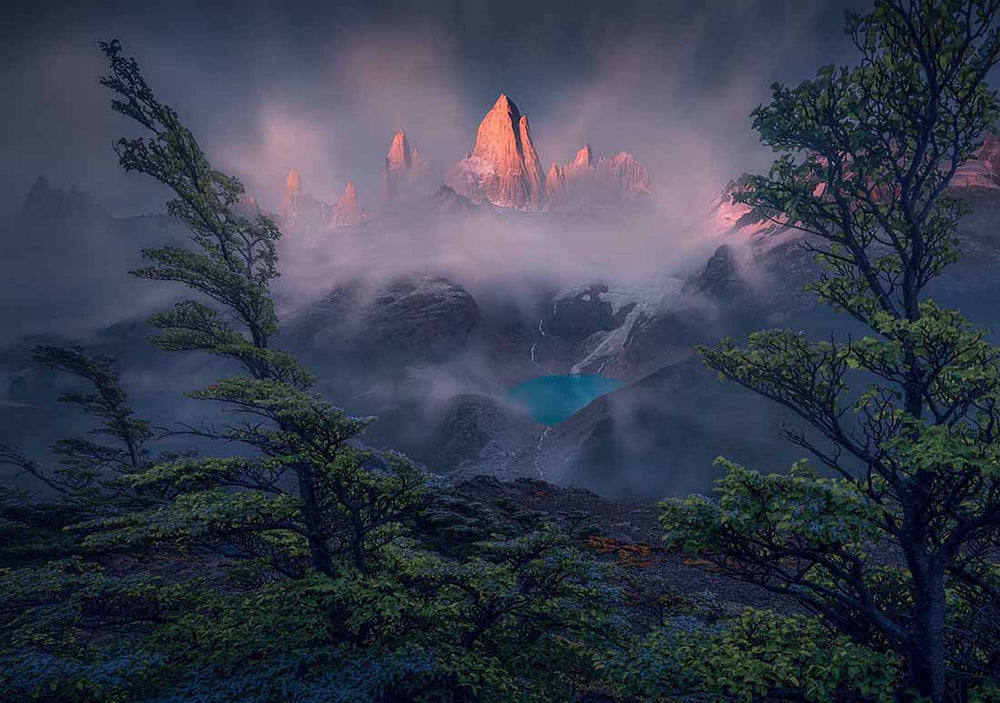 tall green trees frame sharp and pink mountain peaks. The mountain is shrouded in mist and below it sits a glacier and lake with waterfalls either side.