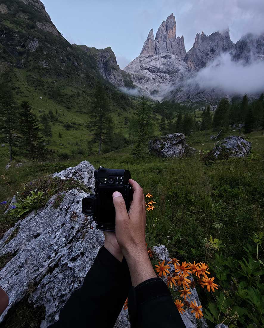 In the foreground orange flowers bloom beside a grey rock. Hands using the rock to balance hold a camera to shoot the scene. In the distance sharp mountain peaks reveal through the low clouds.