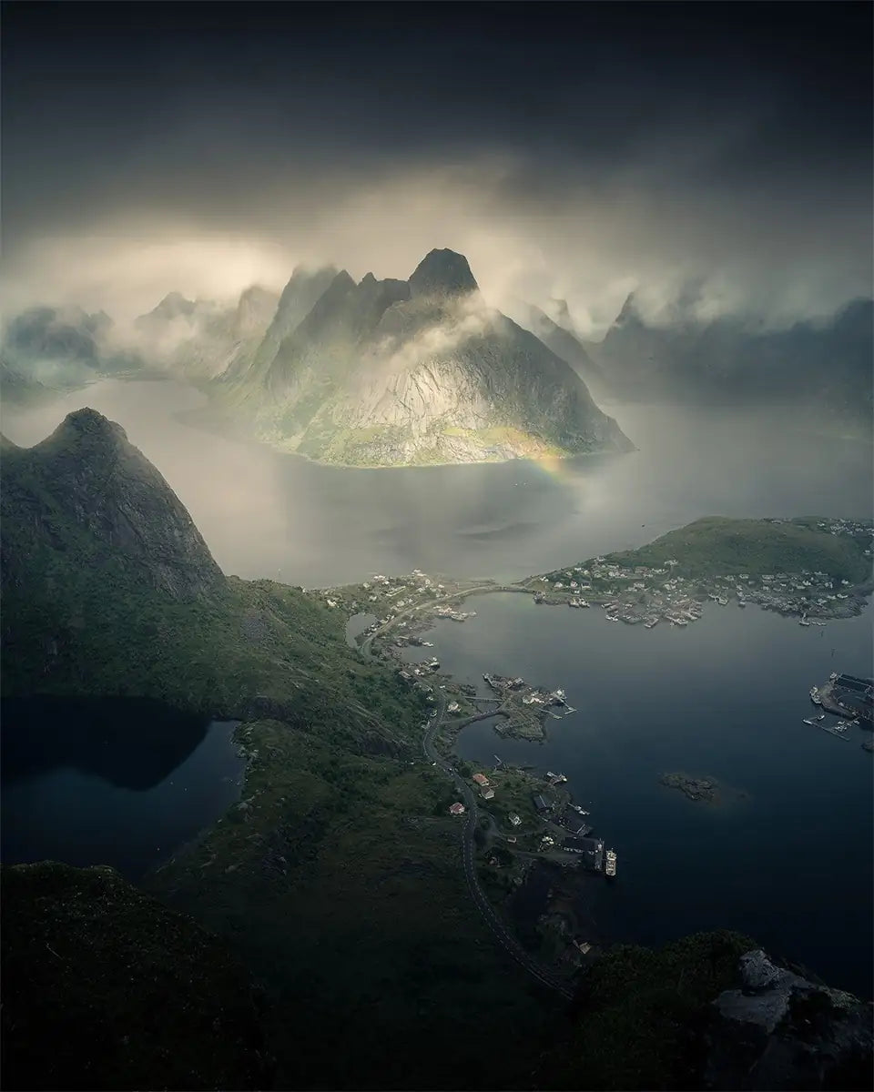 An aerial view of Reinebringen in Lofoten, taken during a cloudy and misty day with the peaks revealing slowly and mist around the tops of them, below the fjords are a glassy dark blue and grey.