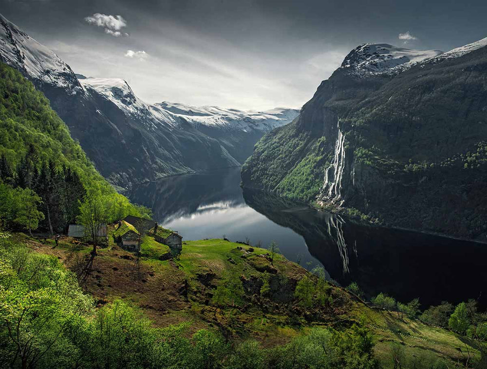 Skageflå in Geirangerfjord during spring time. The farmland is a vibrant green surrounding the other farm houses. the fjord below shines brightly reflecting the seven sisters waterfall.