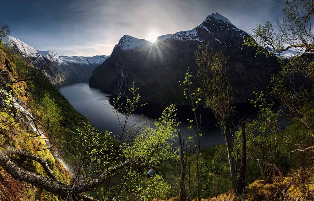  Geirangerfjord during spring time overlooking the seven sisters waterfall from across the fjord. Downy birch trees fill the foreground and a sunstar appears over the snow capped peaks.