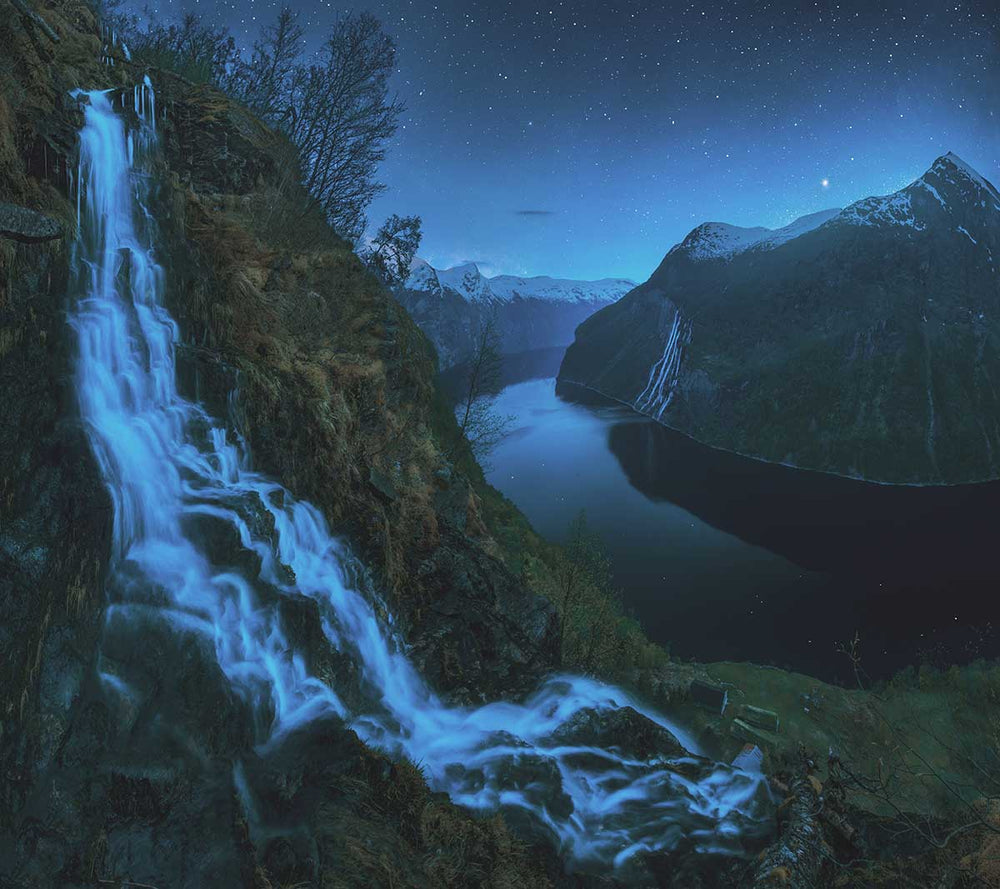 Geirangerfjord at night time. A giant waterfall cascades down a mountainside in the foreground. over the fjord is the seven sisters waterfall and snow capped peaks. stars shine in the night sky.