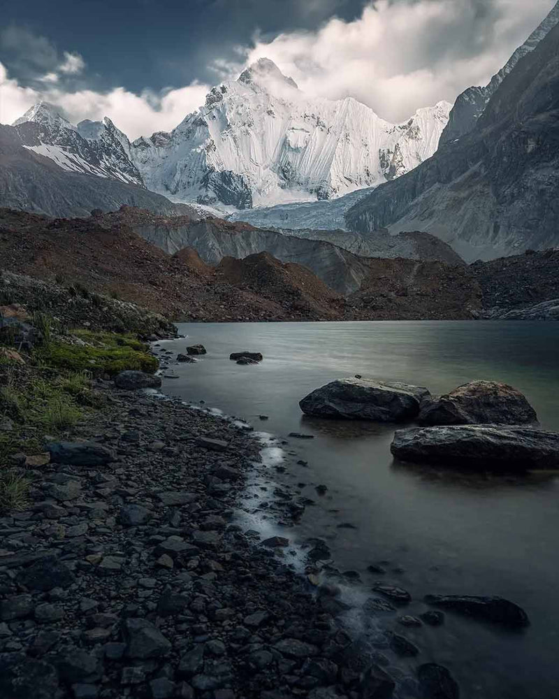 A pebble-covered lakeshore beneath snow-covered Cordillera Huayhuash mountains.