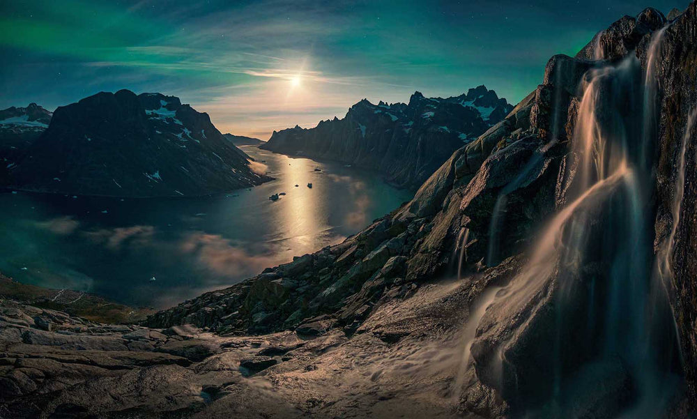 An aurora in Greenland over the fjords. The moon shines brightly in the middle of the mountains. a waterfall frames the scene and enhances the winter mood.