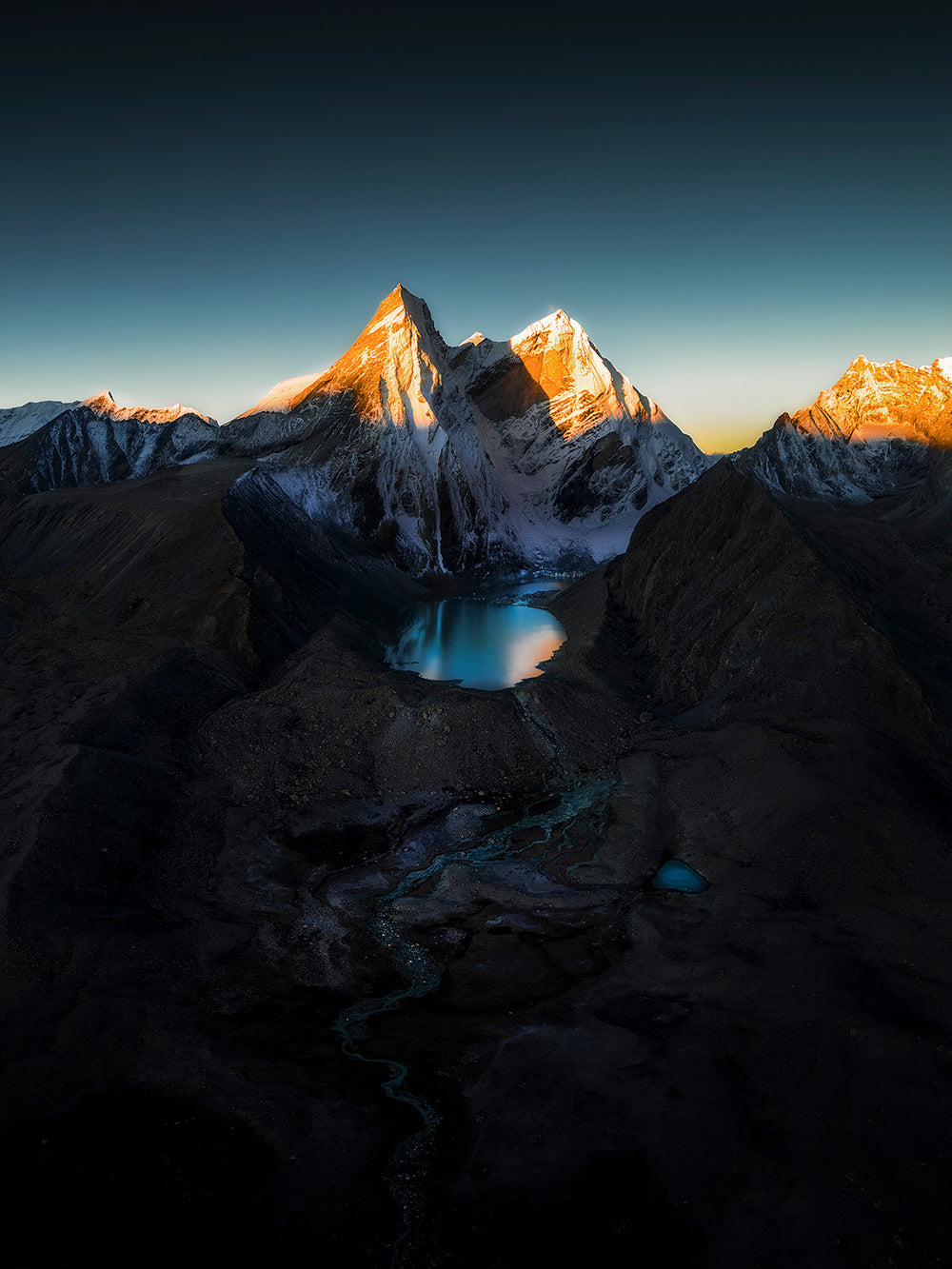 The Himalayas in Tibet with dramatic snowy mountain peaks under a mostly clear sky, a large glacier lake sits beneath the peaks at sunrise.