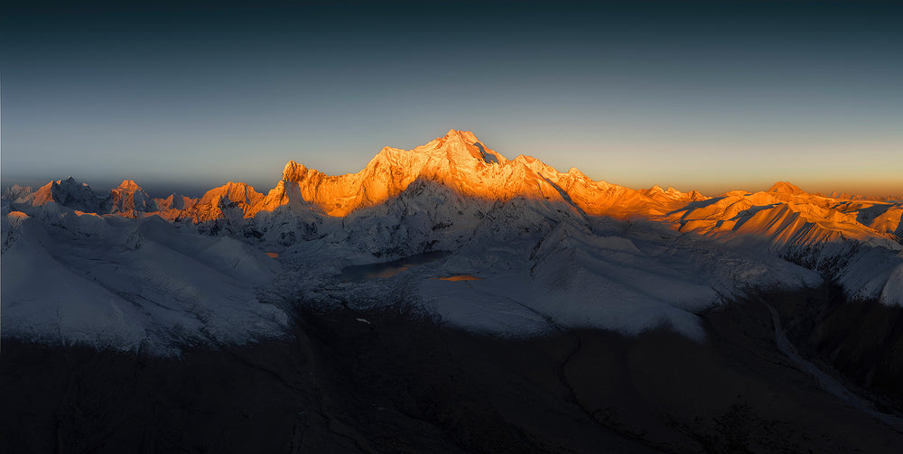 A panorama of the Himalayas in Tibet at sunrise where the tops of the snowy peaks are illuminated with a golden orange light , there is a cloudless dark sky above.