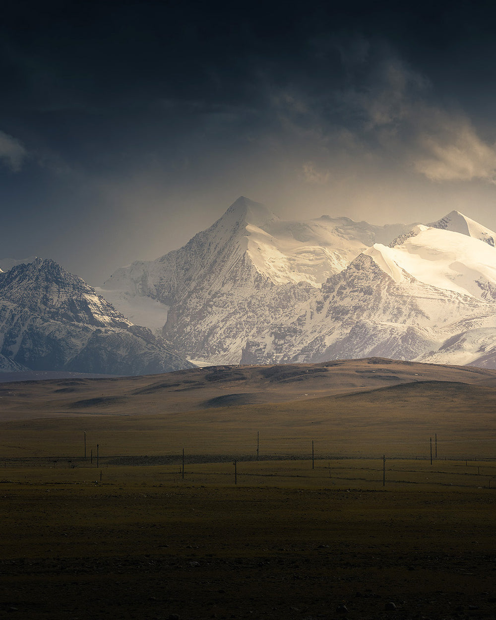 The Himalayas in Tibet with giant snow covered mountains under a dark moody sky, the midground is a green sloping hill underneath the peaks.