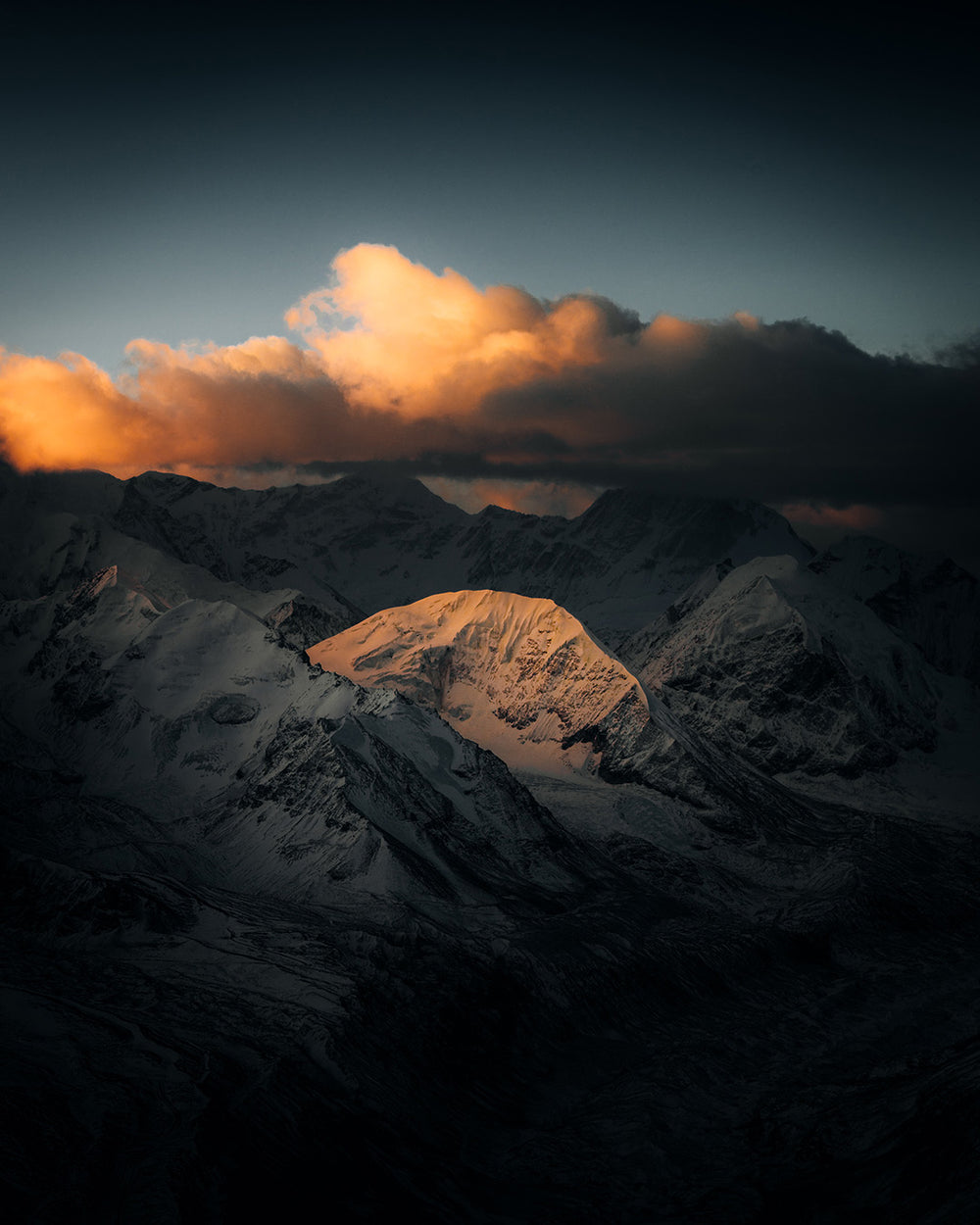 A dramatic landscape in the Himalayas of Tibet with giant snow covered peaks, orange light illuminates the middle peak and orange glow tints the clouds above.