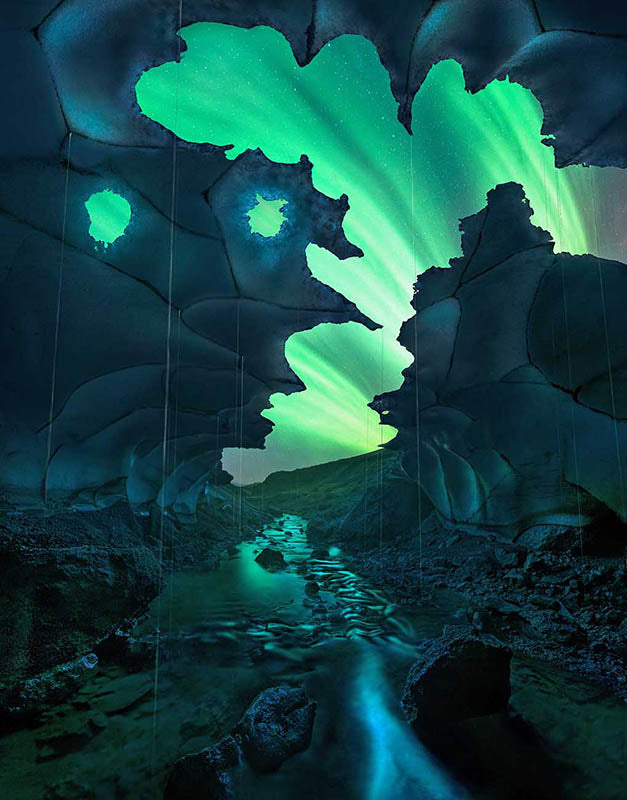 A bright aurora shines through the holes in an ice cave in Iceland during winter. Cracks in the snow create patterns in the ice and a stream run throughs the centre of the cave towards the aurora.