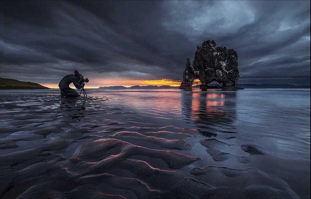 The coast in Iceland during a sunset. Sand patterns create leading lines in the foreground. A large rock formation is in the centre of the frame and through the holes in the rocks the orange glowing sun shines.
