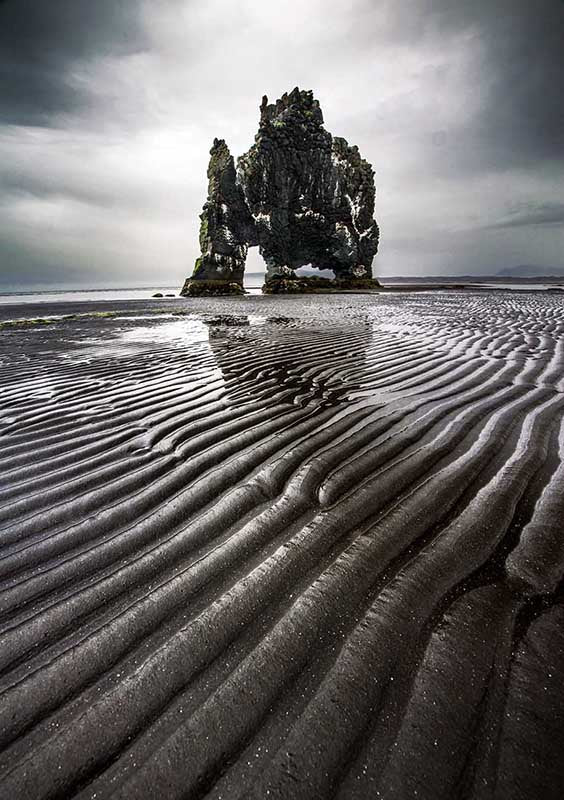 Lines of sand formed by the waves of the ocean stretch long across the beach in Iceland. giant forest covered rock stands tall in the stormy light, carved by the waves over time it has holes in the bottom.