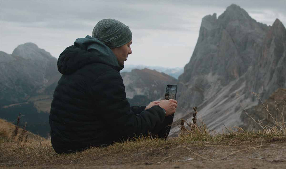 a man sits on the side of a mountain holding a phone to some flowers. Mountains in the background.