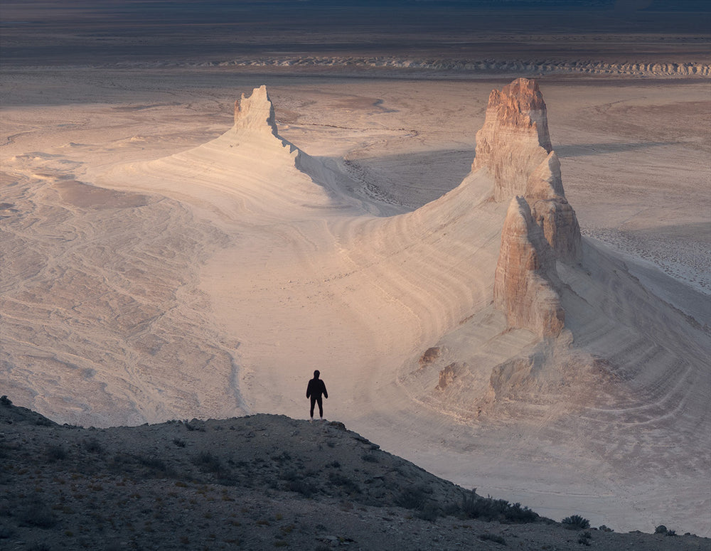 A person stands on the edge of a cliff at sunset looking at the vast desert and mountains of Kazakhstan Mangystau.