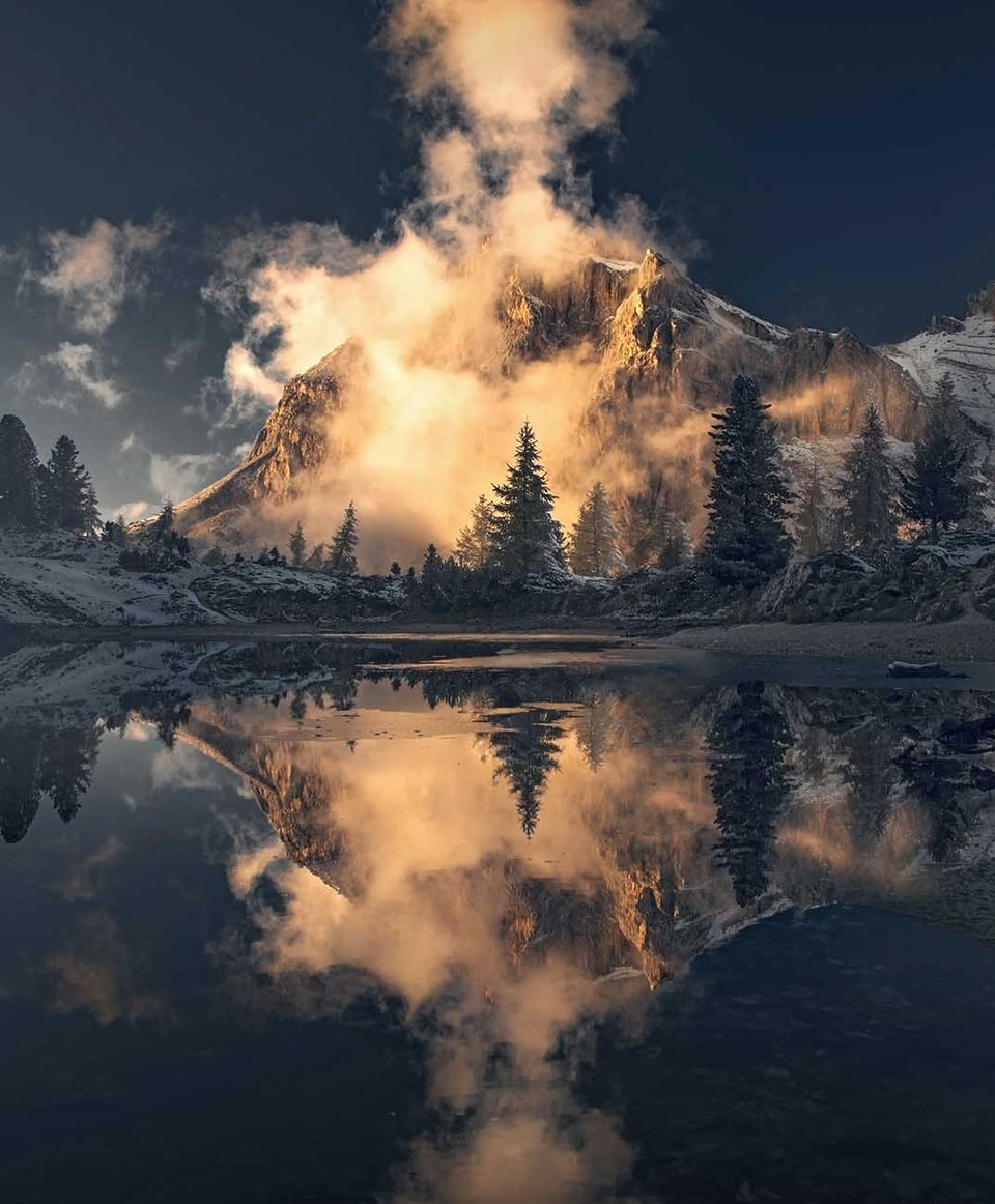 A dramatic mountain landscape in the Dolomites with snow-covered peaks, dark evergreen trees, and the reflection in Lago di Limides. A large glowing cloud rises from the mountain and is mirrored perfectly in the water.