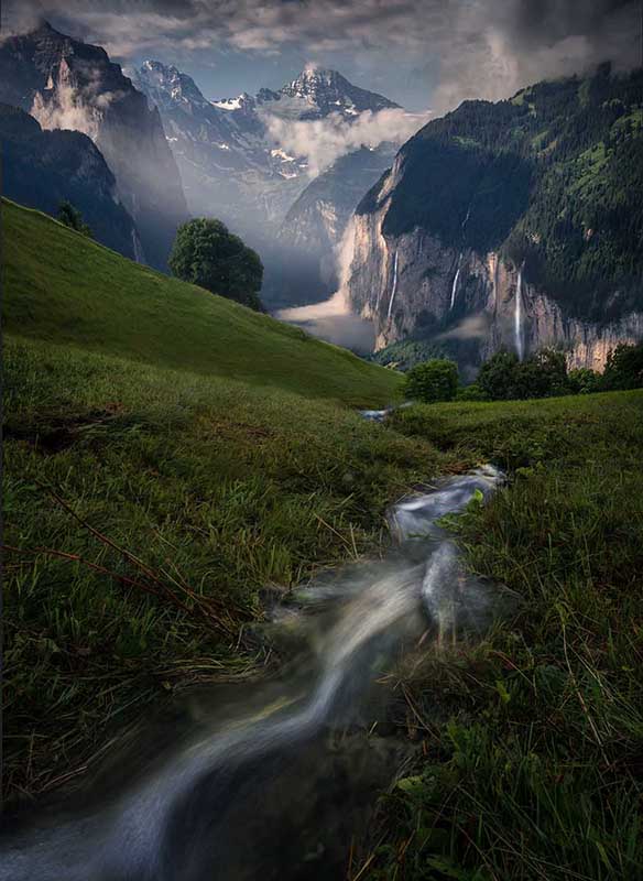 a stream runs down a green valley to mountains beyond. The mountains have waterfalls running down the cliff face, buchenbachfall, spissbachfall nd staubbachfall.
