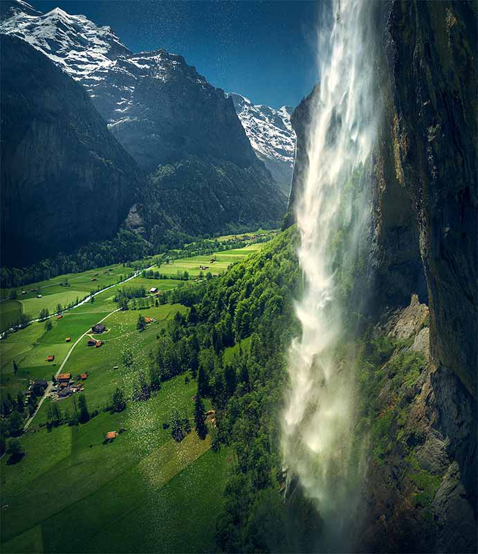 A giant waterfall cascades off the side off a cliff face into the green valley of lauterbrunnen village below in summer.