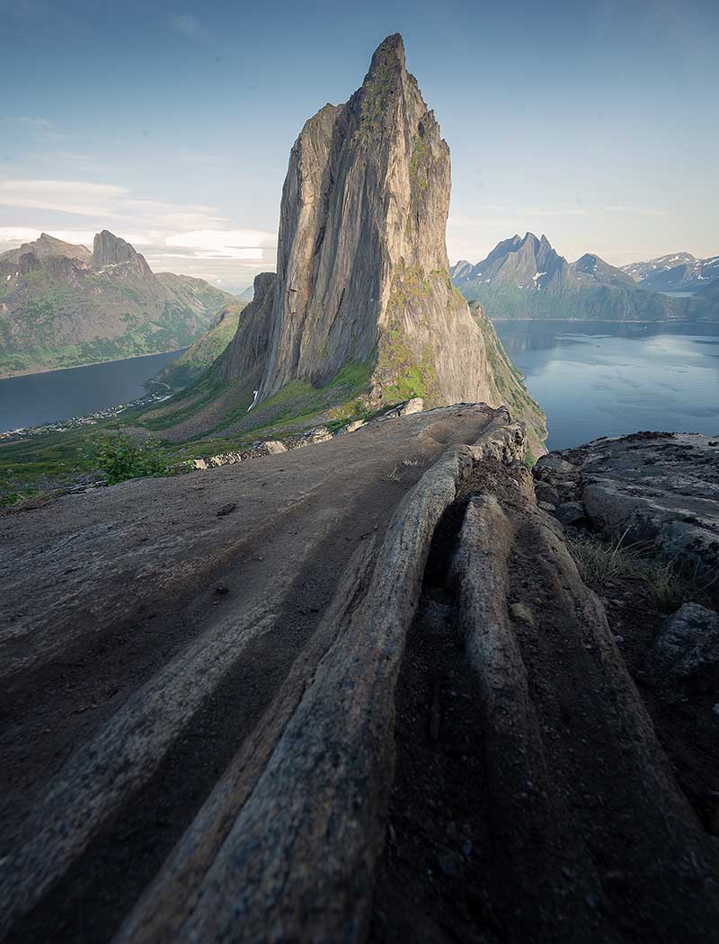 Cracks and crevices on the ridgeline above Segla create leading lines towards the sharp mountain. Below a glassy fjord shines brightly in the midnight sun.
