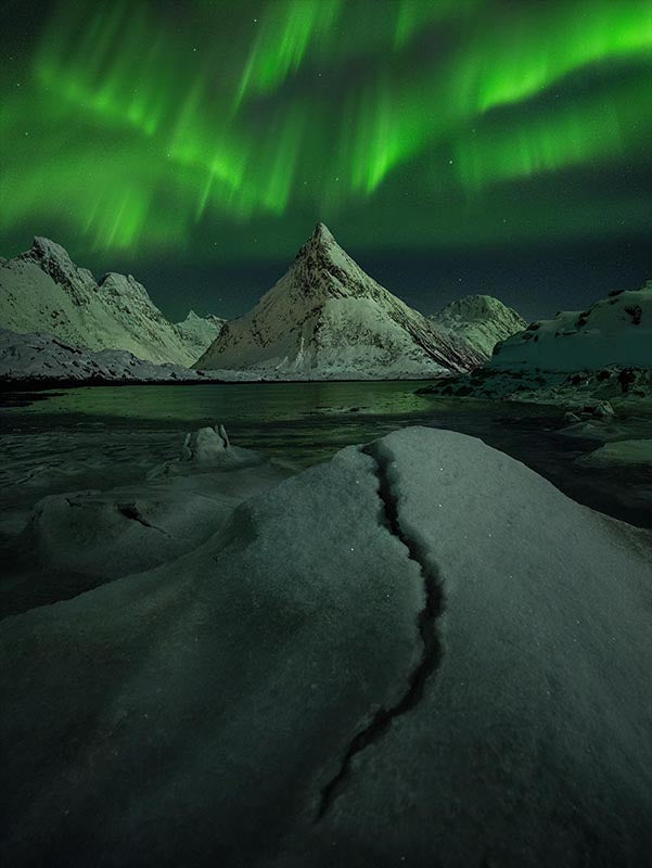 luminescent green norther lights above a pointy snow covered mountain in lofoten. In the foreground a large sparkly ice crack shows in the fjords. 