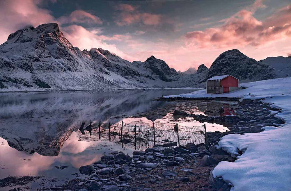 A red fisherman's cabin stands alone by a snow fjord with a pink sunrise reflecting the mountains.