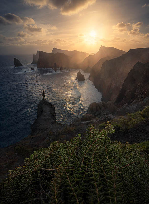 A person stands on the edge of a rocky out cliff looking into the sun on the coast of madeira, Portugal. the sunrise is golden and lights up the white capped ocean and layers of cliffs.