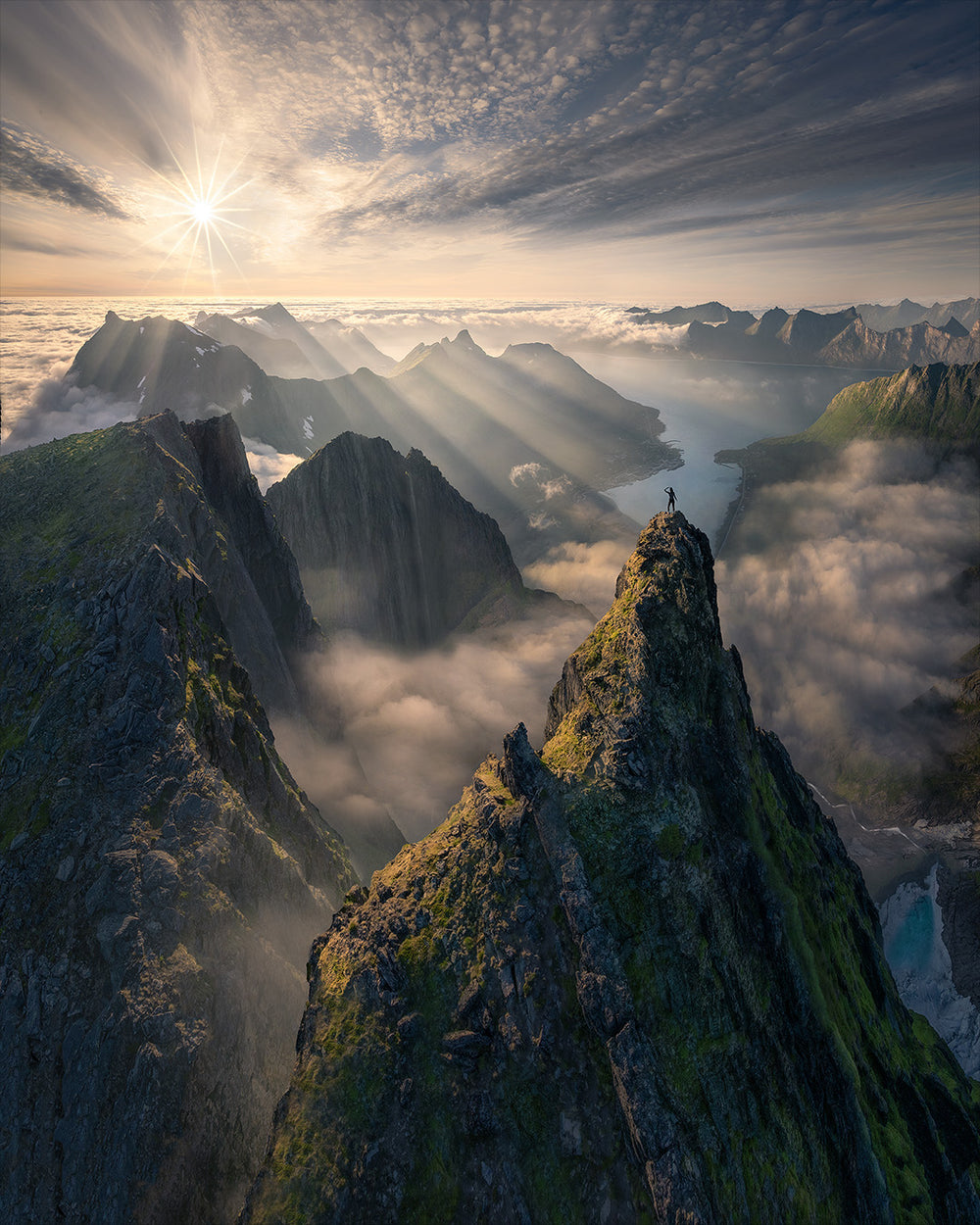 Midnight sun in Senja at Roalden viewpoint. Madison McCardle stands on top of a rocky ridgeline looking at the and fjords below. Sea fog above the fjords and below the mountains is illuminated in a golden glow. Light rays pour across the top of the fjords.