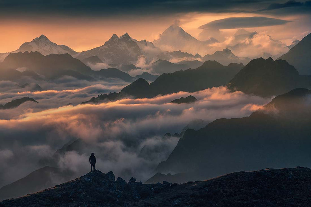 a man stands above the clouds looking at the layers of Himalayan mountains during a pink sunrise.