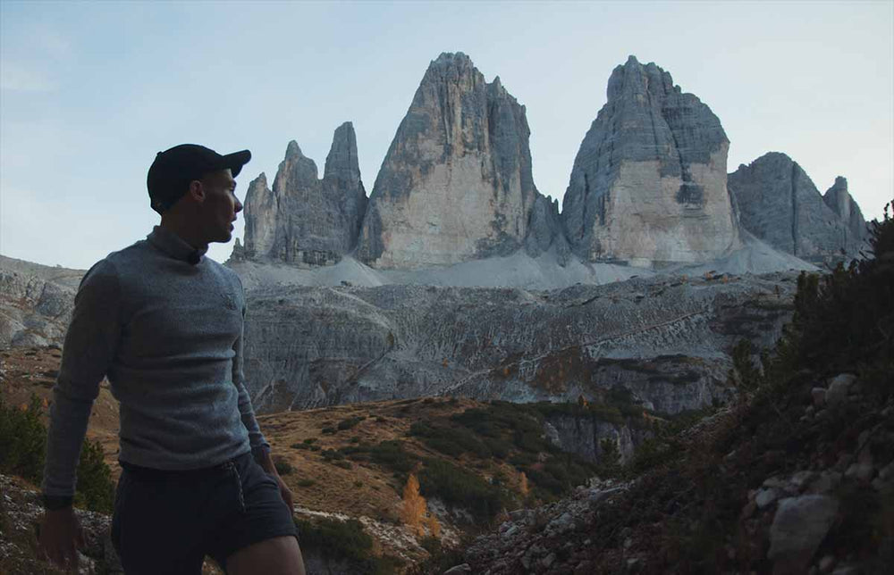A sexy man stands in front of tall mountain peaks in the Dolomites.