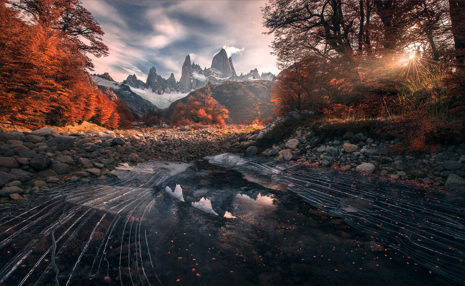Mount Fitz Roy's jagged mountain peaks above a rocky lake, framed by brilliant orange autumn trees with sunlight breaking through the right side of the canopy, the frozen lake has cracked lines on the surface.