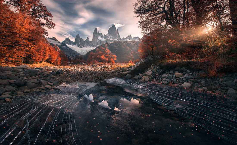 sunrise over mount fitz roy in autumn. Leading lines in a frozen ice lake point to the mountains between the sunrise and a forest of red coloured trees.
