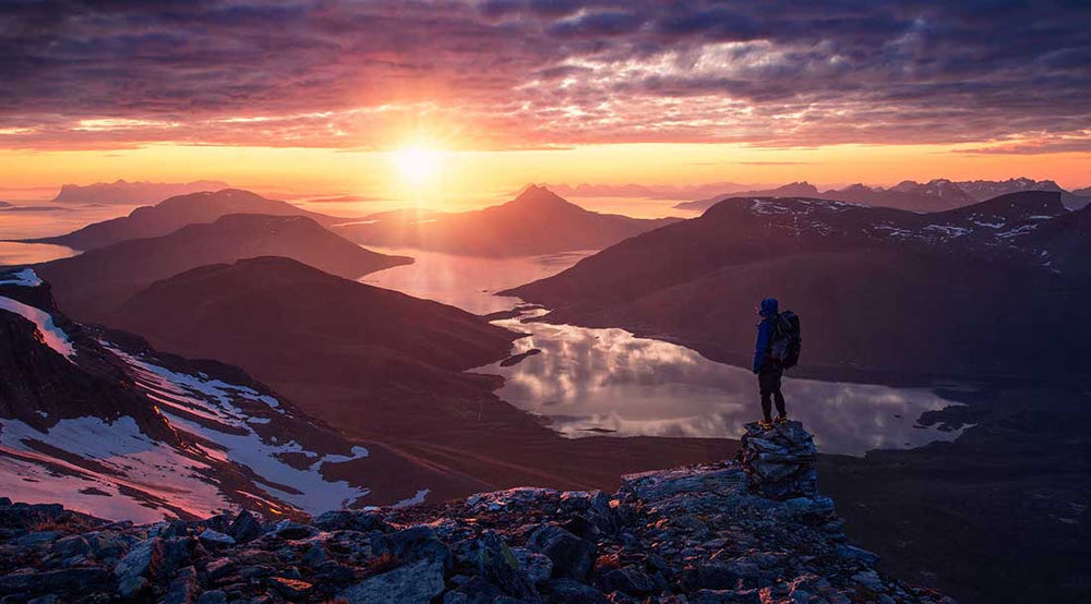 The silhouette of a man wearing a backpack standing on the edge of a ridgeline in northern Norway during midnight sun. The sun casts an orange glow into the clouds and over the fjords below.