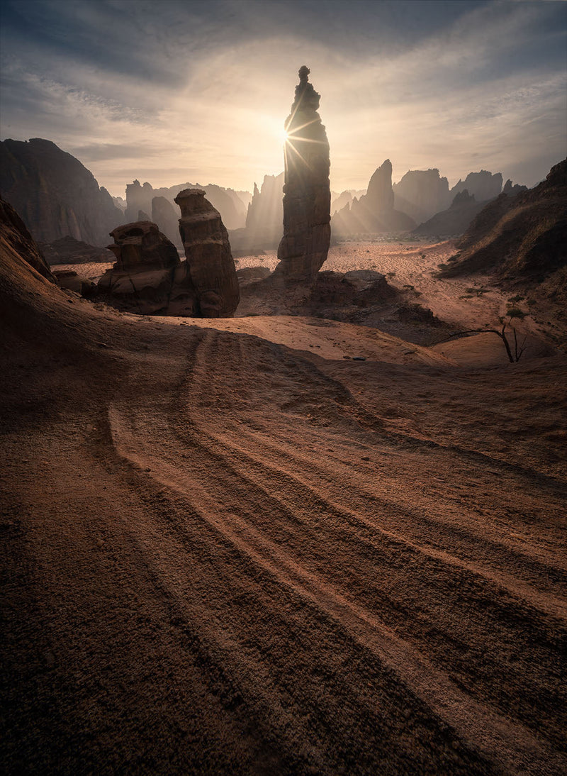 Sunlight behind a slender sandstone spire, with curved erosion lines across the Saudi desert foreground.