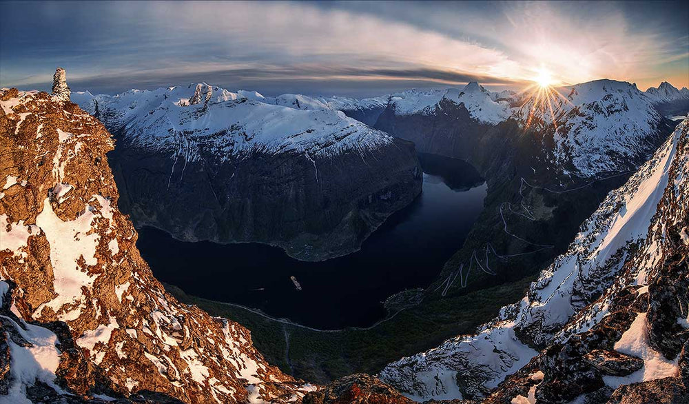  Geiranger fjord in Norway taken in spring taken from the top of a high viewpoint looking down at the fjord and winding road beneath it. A stunstar is in the far right of the mountains. The mountains are snow covered.