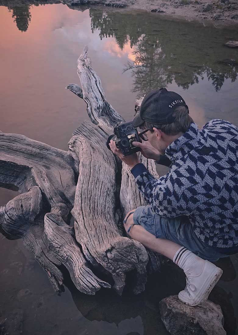 Max rive uses his knee as a tripod to balance his camera. The foreground is a dead tree trunk floating in a lake. The whole scene is illuminated pink from the sunset.