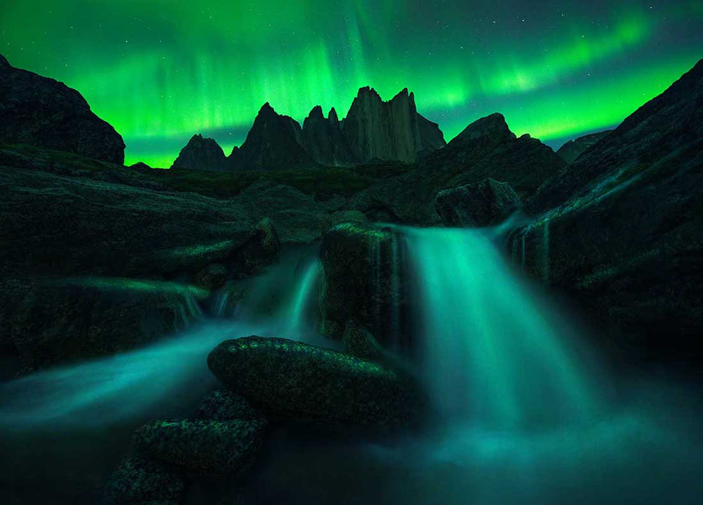 brilliant green northern lights stretch across the horizon above a giant mountain and waterfall at night in Greenland.