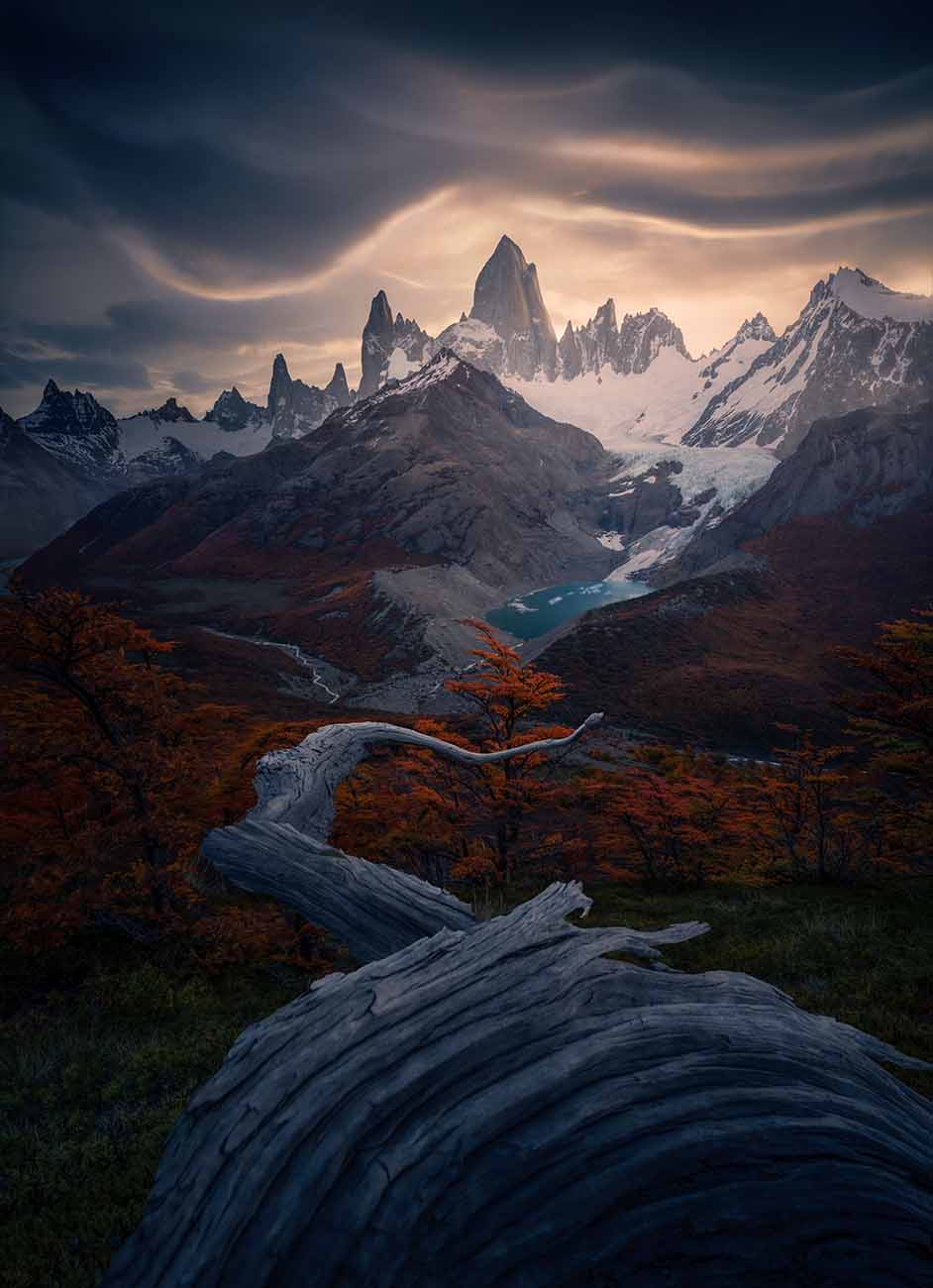 A dramatic mountain landscape in Patagonia featuring jagged snow-capped peaks under a cloudy sky with warm light, a blue glacier lake in the valley beneath the peaks, and autumn-colored trees with a large weathered log in the foreground.