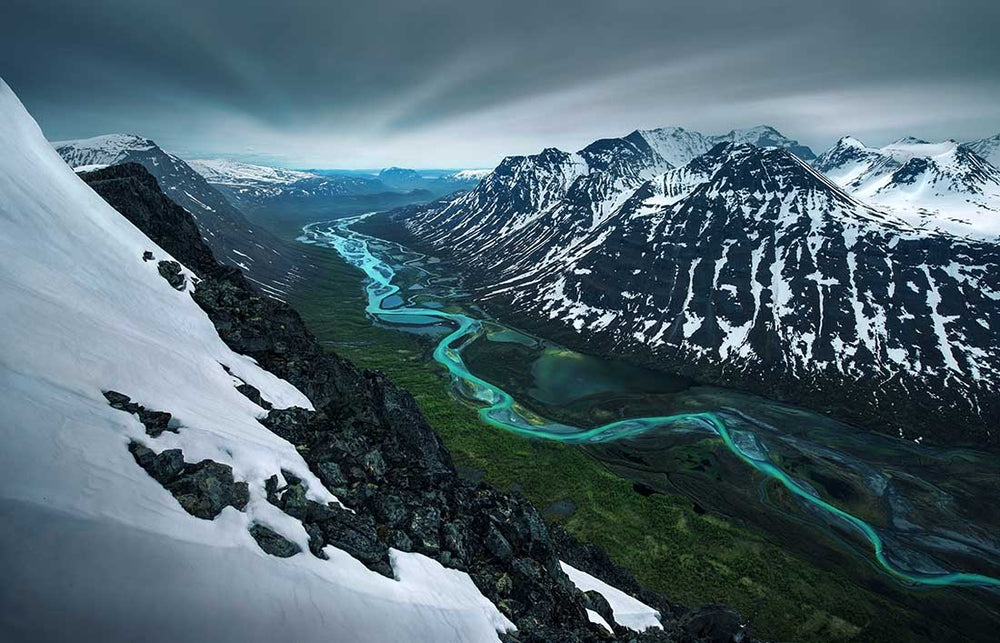 A photo taken by Max Rive during the Midnight Sun in Sarek, Sweden. An ultra blue and green glacier river flows between the snowy mountains and is surrounded by green forest.