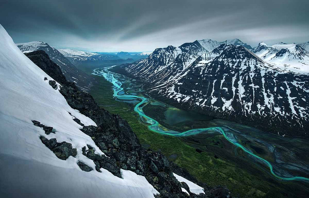 Sarek in Sweden in Spring, taken by drone high above a glacier river which winds through a valley surrounded by snow covered mountains.