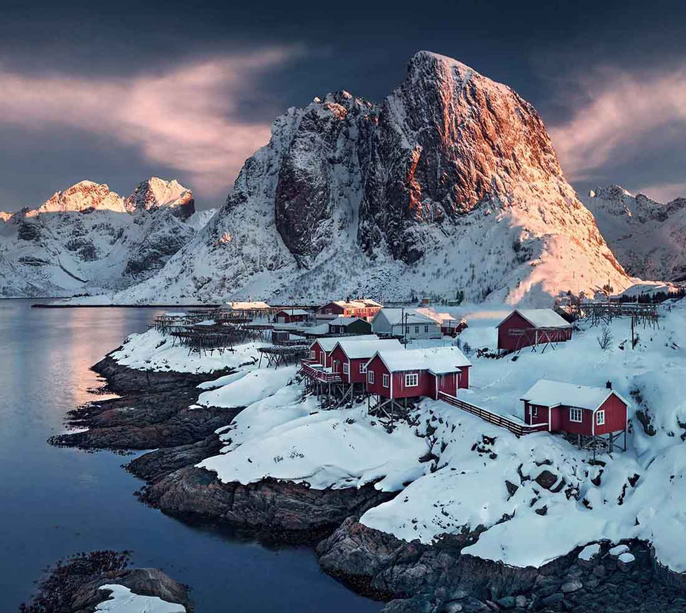 Red fisherman cabins in winter snow stand by a fjord and underneath a giant mountain in a pink sunrise.