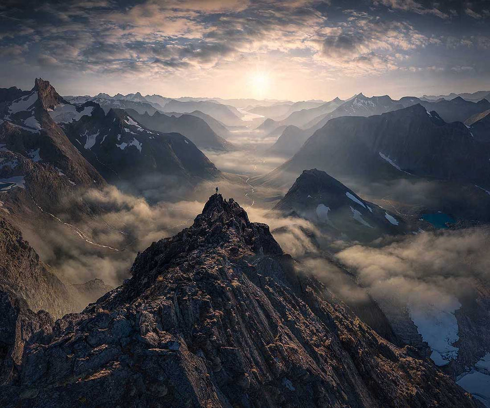 A six shot panorama of romsdal mountains in sunset Norway by drone. the silhouette of a man standing on the center mountain looking at the fjords below.