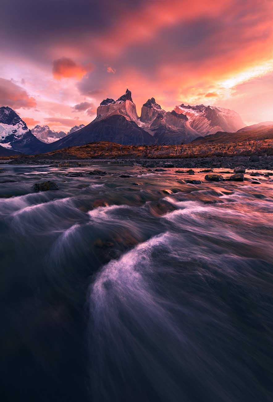 A dramatic landscape the impressive mountain peaks of Cordillera del Paine under a vivid pink and orange sky, with flowing water in the foreground reflecting pink, orange, and purple light.