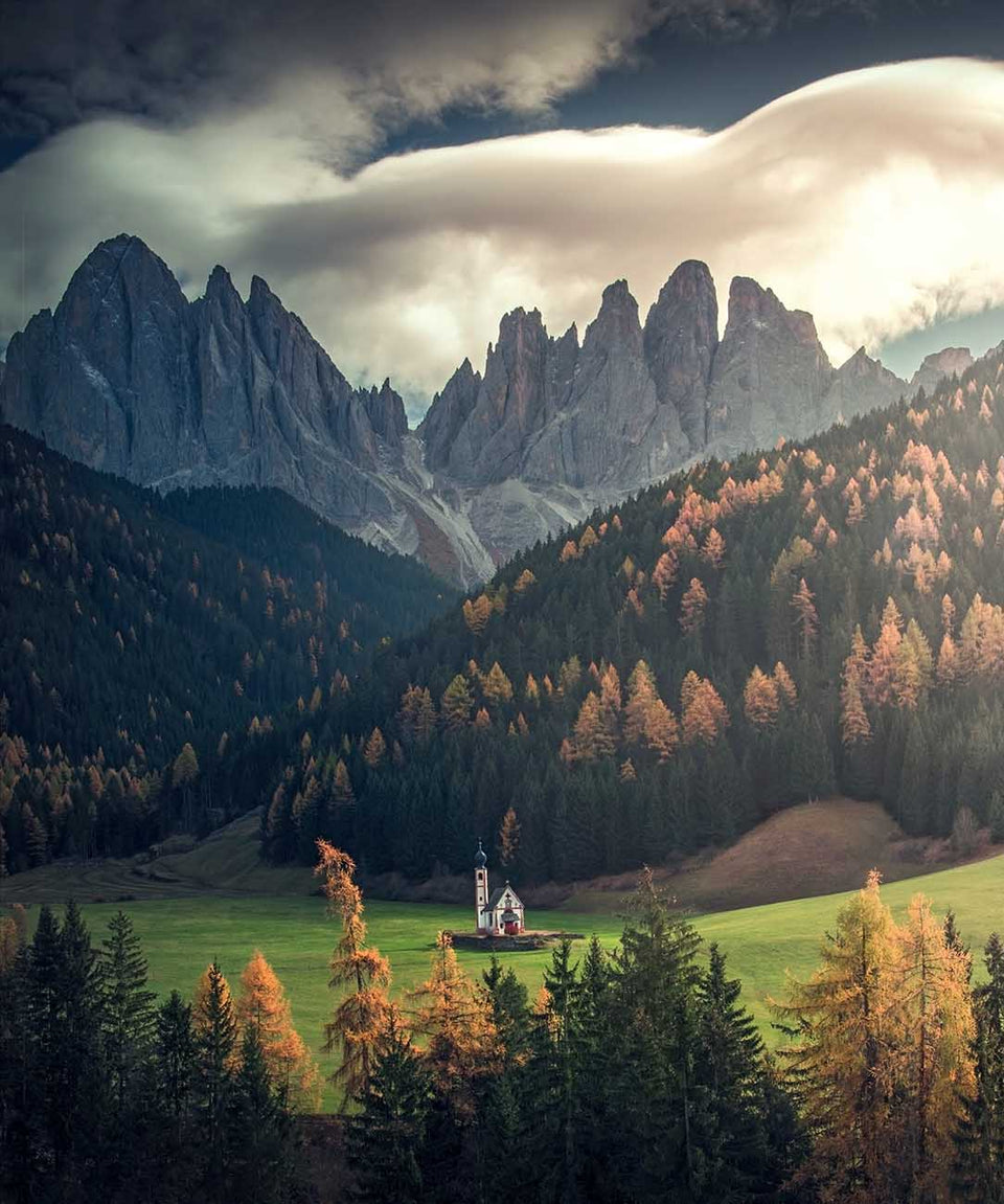 St Johann Church in the Dolomites in a green valley surrounded by evergreen forests, golden autumn trees, beyond are jagged rocky peaks under a cloudy sky.